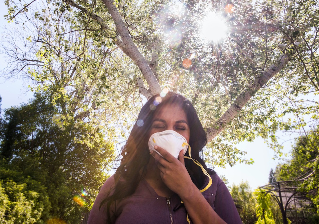 a latina woman holds a mask against her face, sun streams through trees in the background