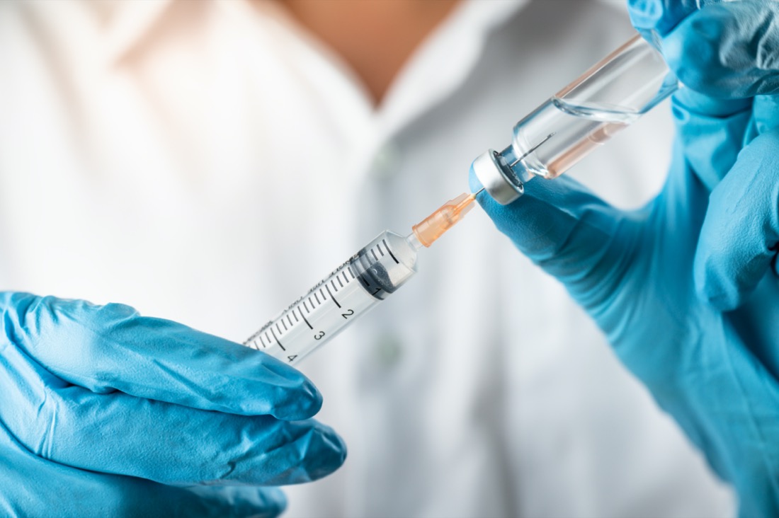 close up shot of a vaccine needle dipping into a vial. the person holding the needle is wearing blue latex gloves and a white shirt