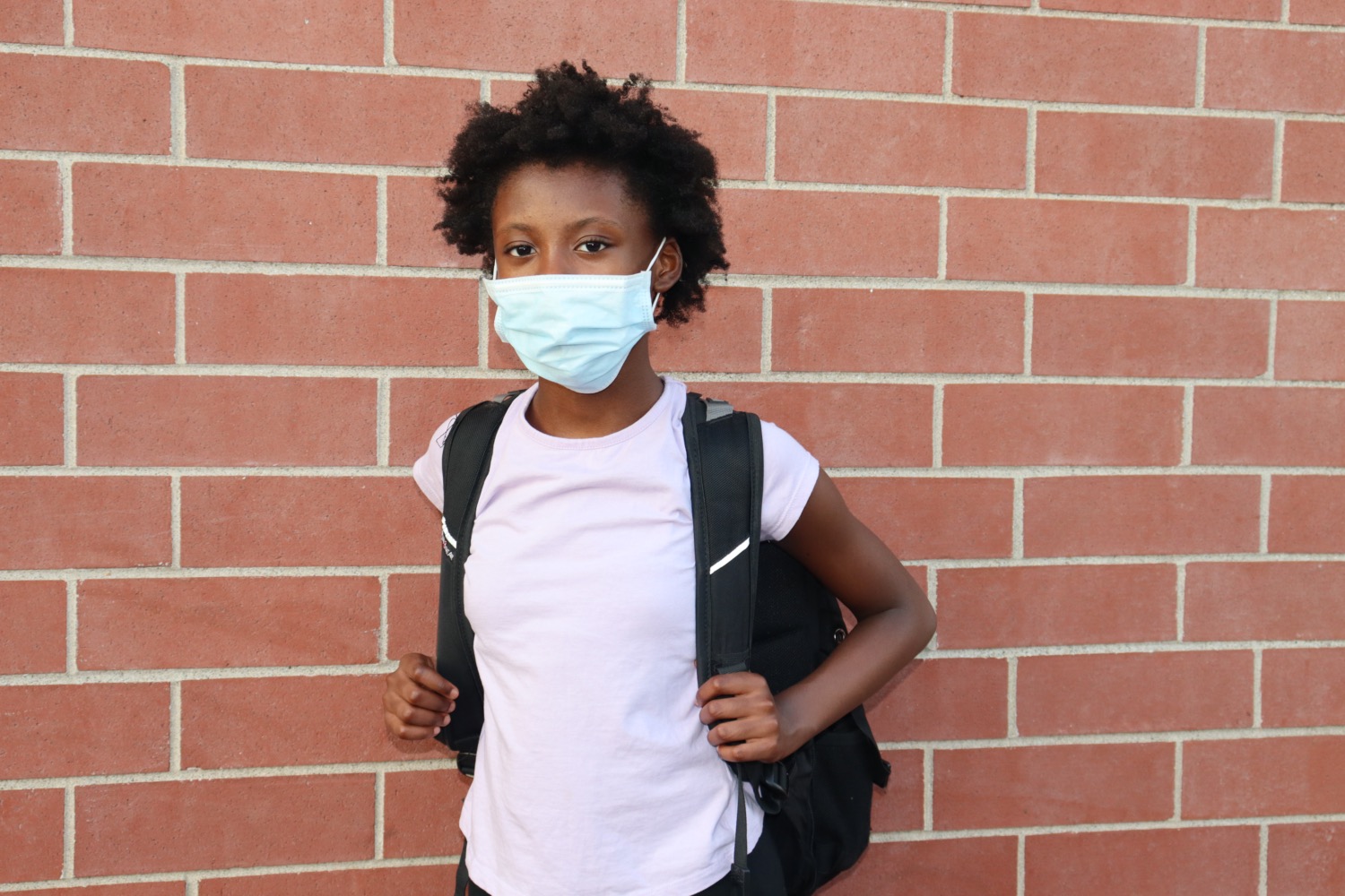 black kid wearing mask stands in front of brick wall with a backpack on