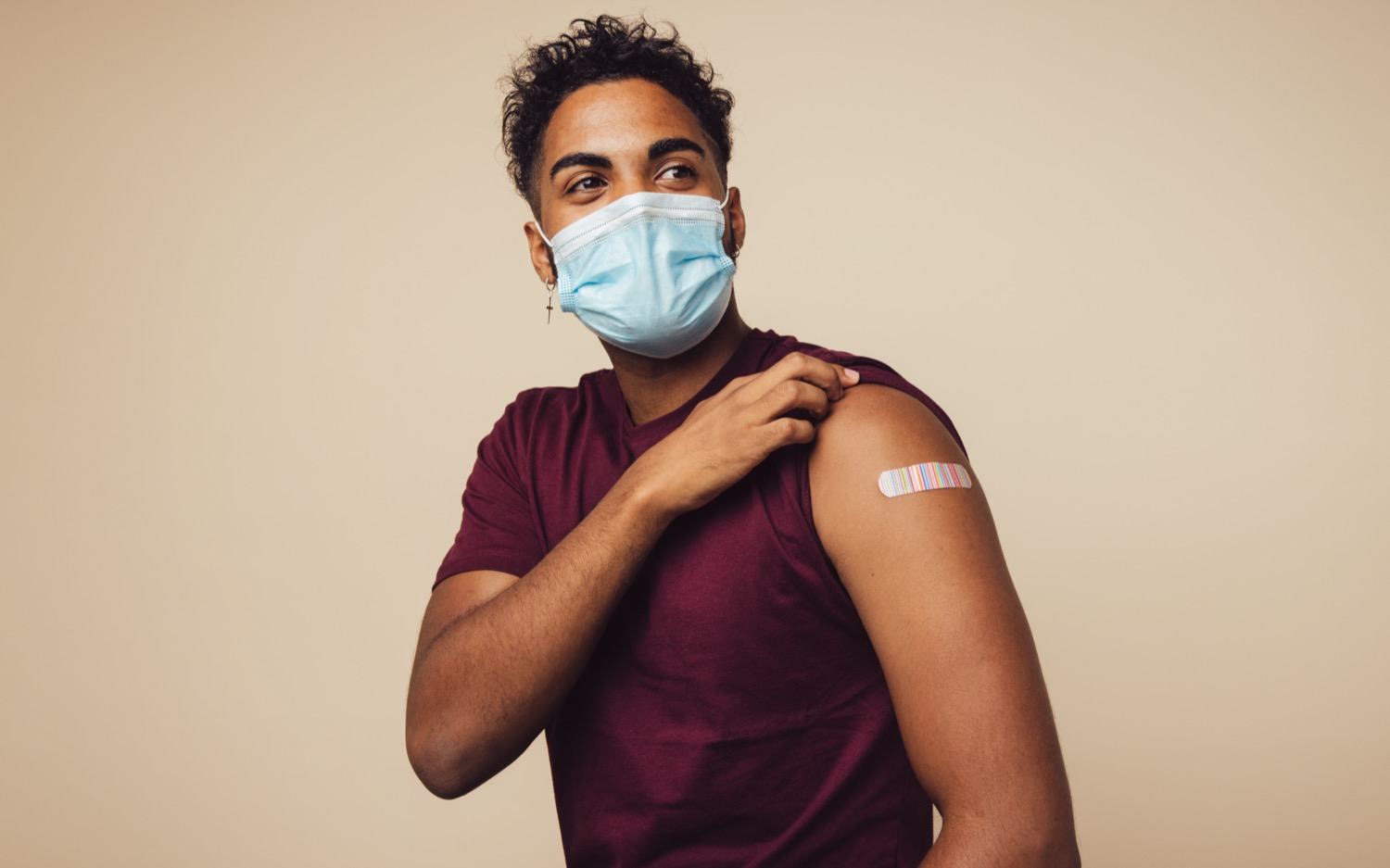 a brown man wearing a mask pulling up his left shift sleeve revealing a bandaid on his arm after a shot