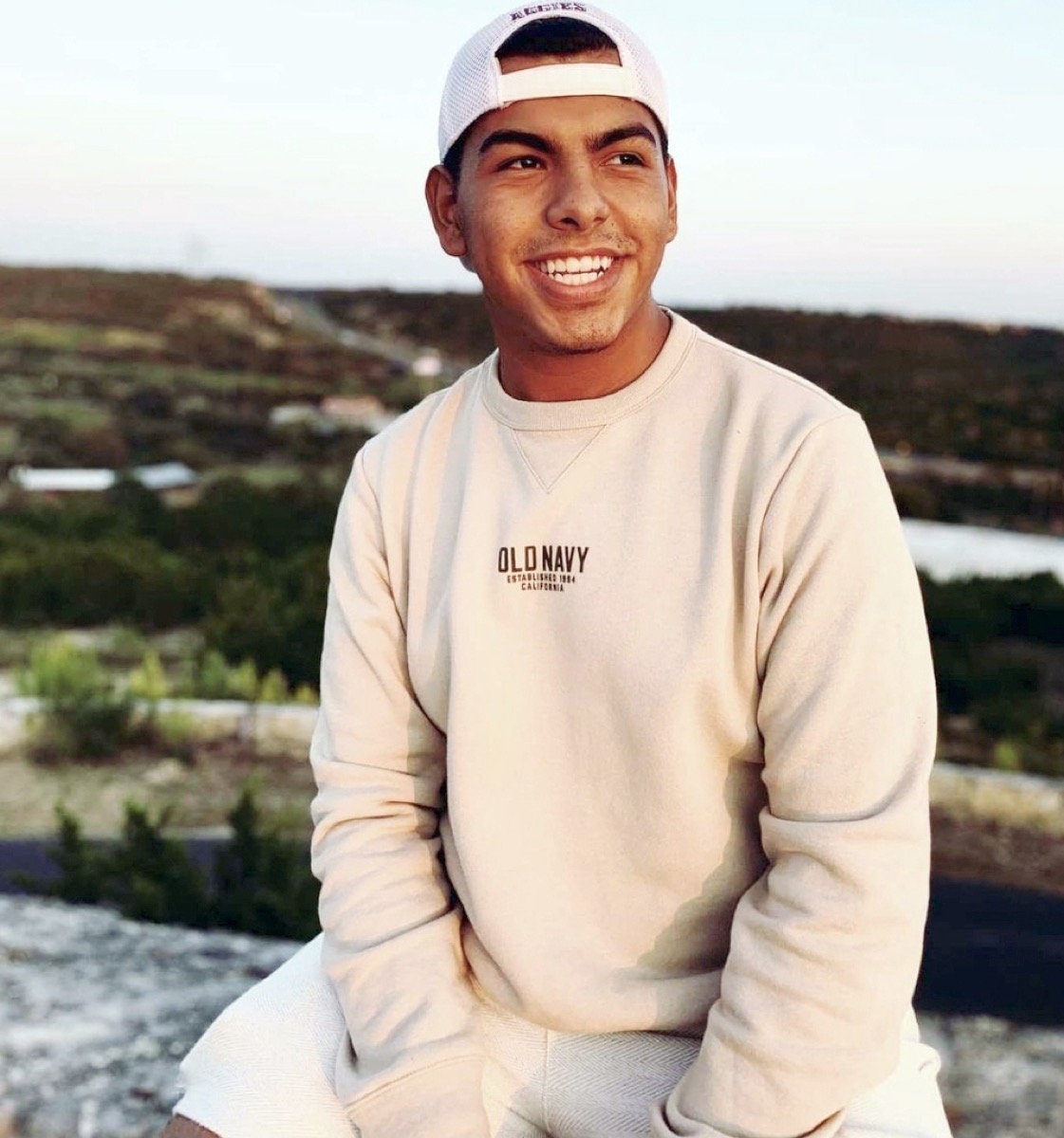 a young hispanic man in a white cap and cream sweater smiles sitting outside