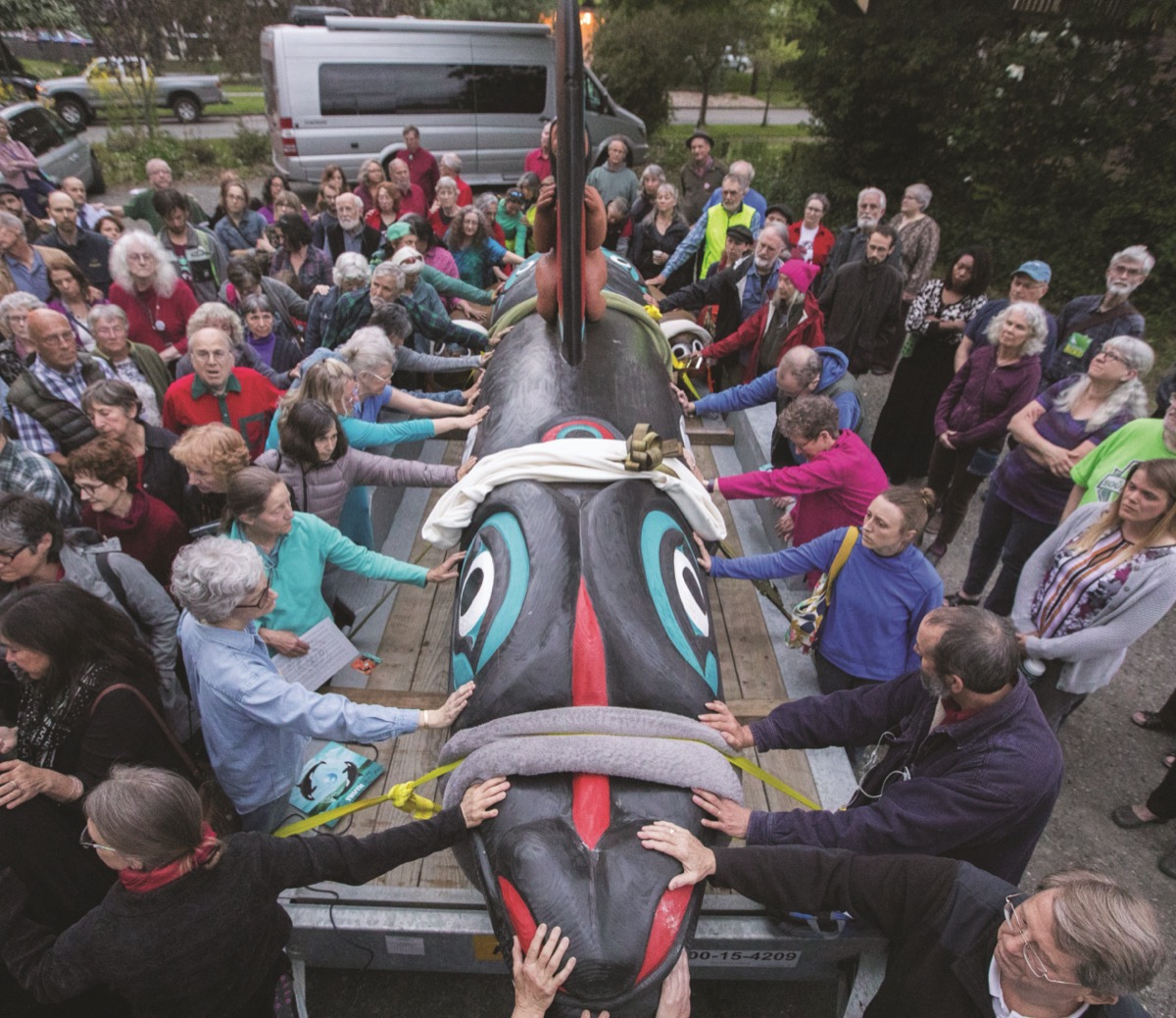 a group of people surround and carry a colorful painted totem pole of an orca