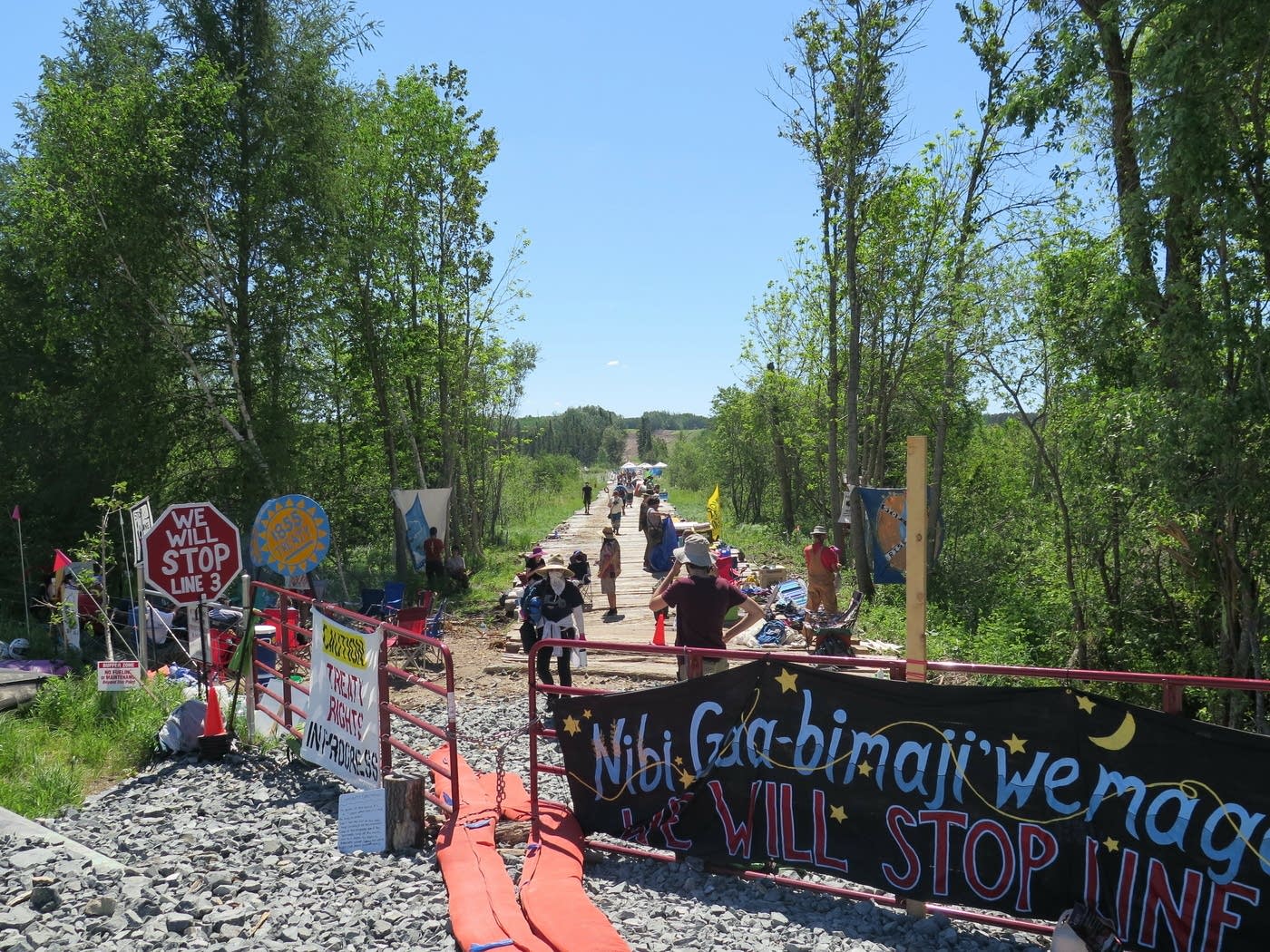 a gravel road that has a gate covered in signs in indigenous languages. signs in english read "we will stop line 3" and similar phrases. beyond the gate on the road protestors line out