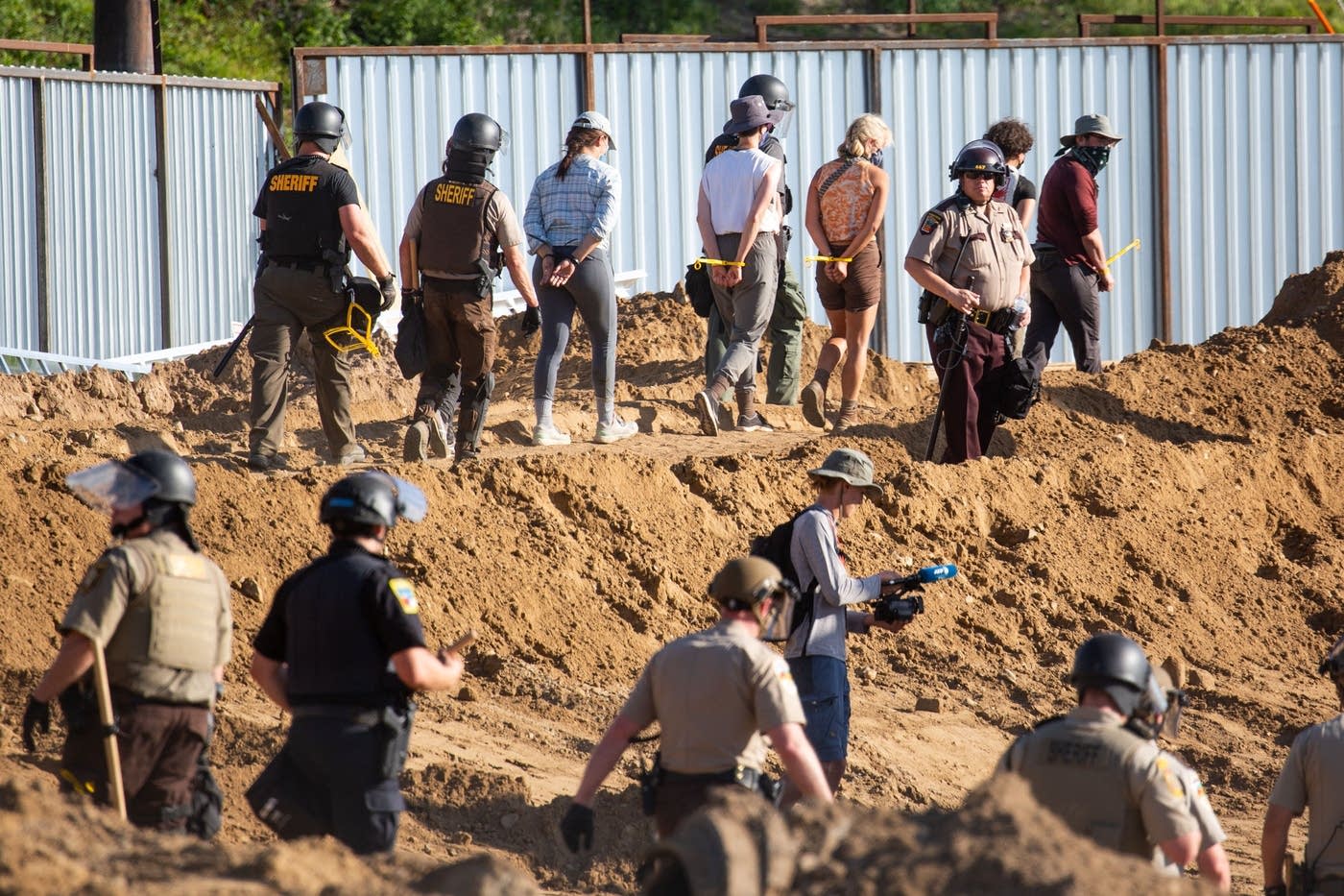 a dirt construction site. a row of protestors handcuffed are being escorted off the property by police officers