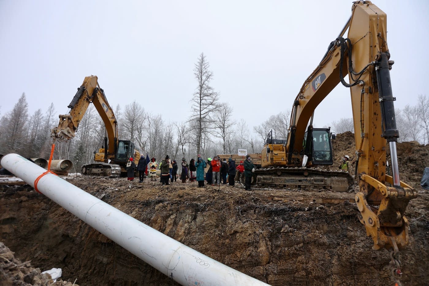 a construction site with two large armed tractors installing a pipeline. between the two tractors is a crowd of protestors.