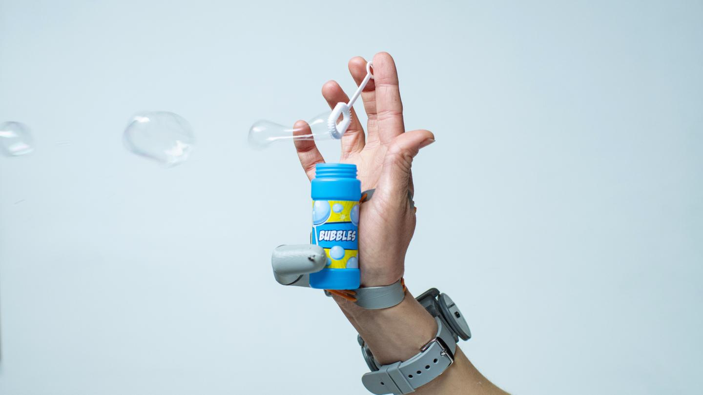 a white hand with the additional thumb prosthetic attached, allowing the user to hold a small bottle of blowing bubbles with the additional thumb while another finger holds the bubble wand