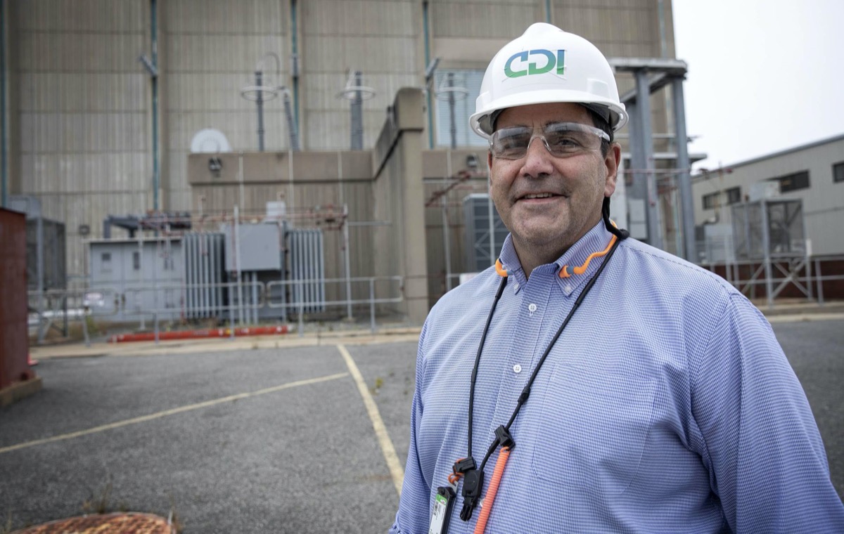 a man in a white hard hat wearing safety glasses and blue shirt stands in front of a power plant