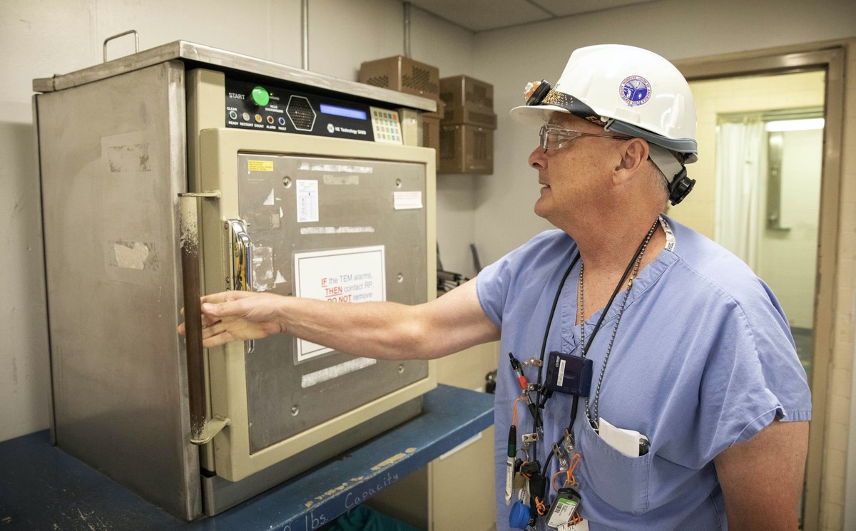 a man in safety glasses and a white hard hat wearing a blue surgical gown opening up a metal box