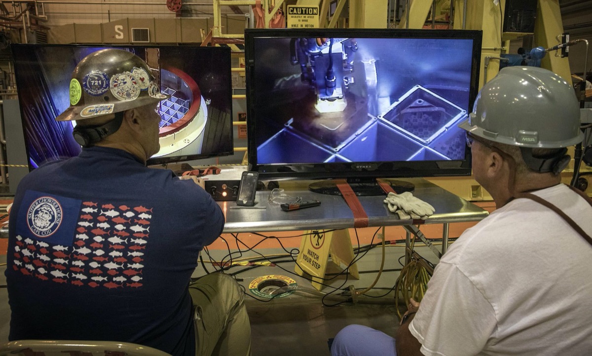 two men in hard hats watch a monitor of equipment being lowered into a fuel pool