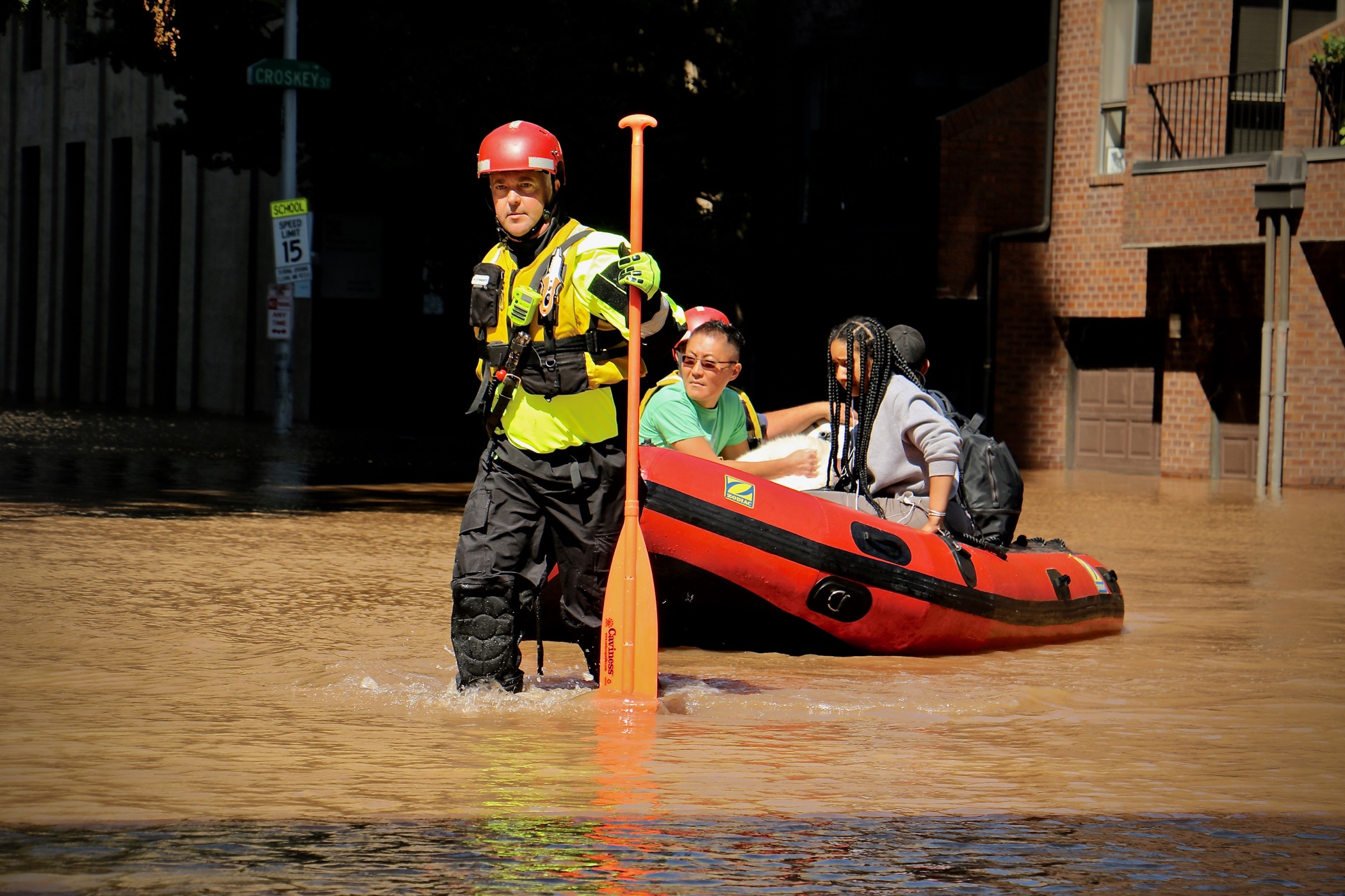 a white man in a hazard vest, hard hat, and water pants holds an oar with one hand and pulls a small inflatable rescue boat with four people in it through a flooded street. the water goes up to his shins