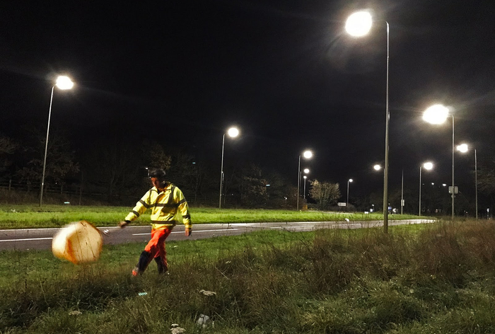 a person in a bright yellow jacket and orange pants swings a bug net along the side of a brightly lit road in the middle of the night