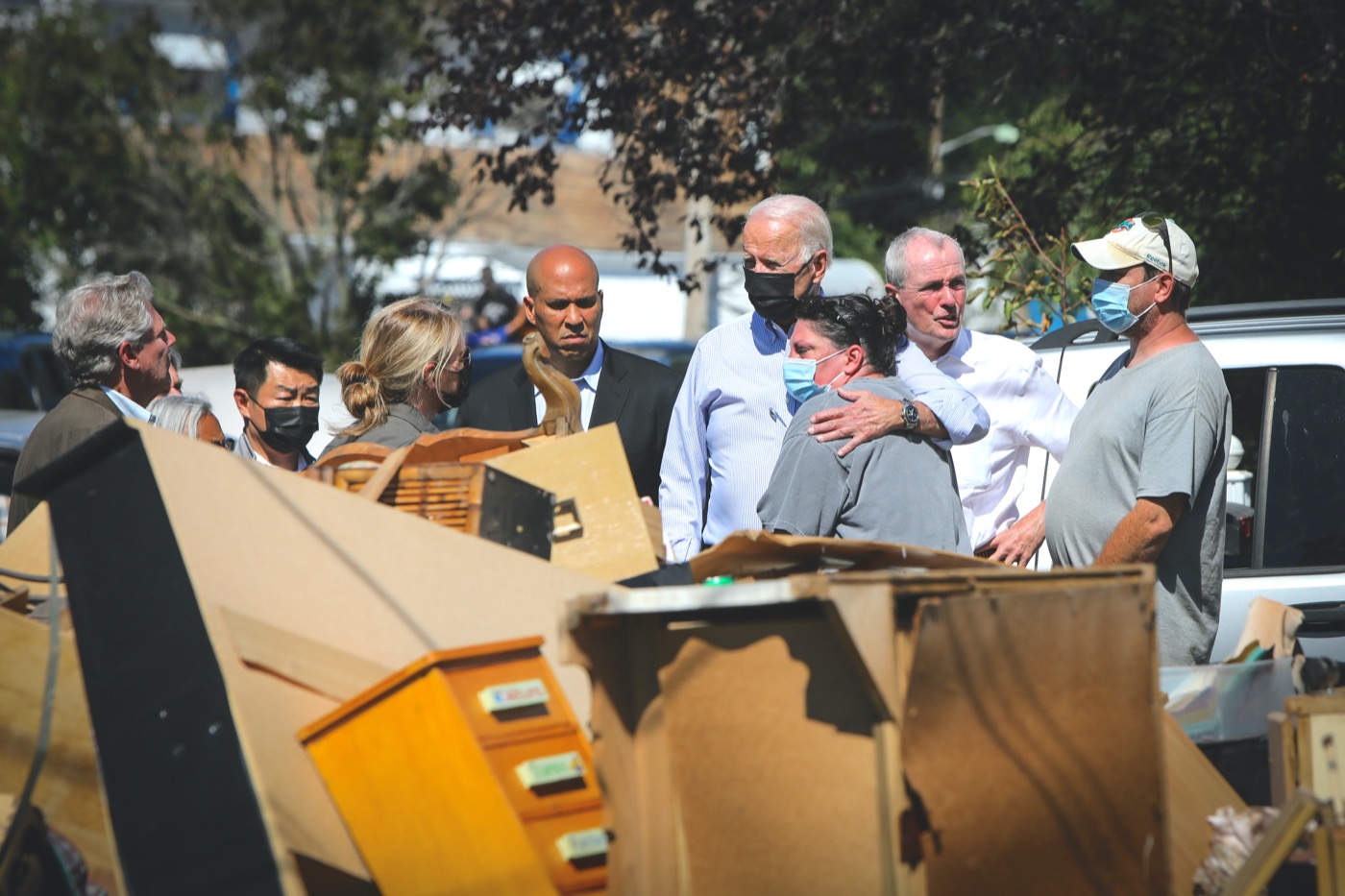 president biden comforts a white woman as they look on to wreckage and debris from hurrican ida. both of them are wearing masks a small group of people stand behind them