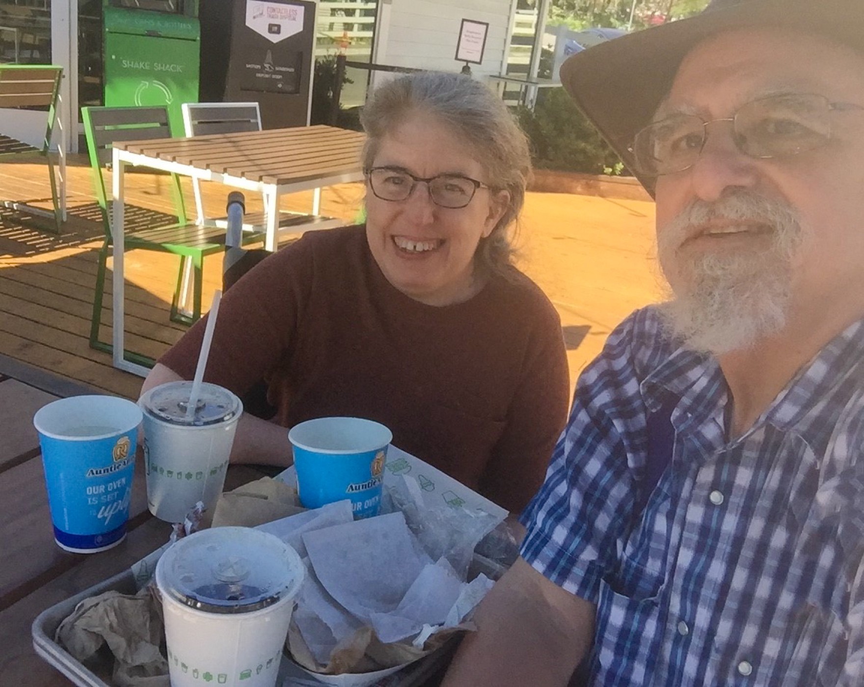 Ingrid and Ken are sitting next to each other, smiling, and looking into the camera. Ingrid is wearing a red shirt and Ken is wearing a blue checkered shirt. On the table are food trays, empty wrappers, and drink cups.