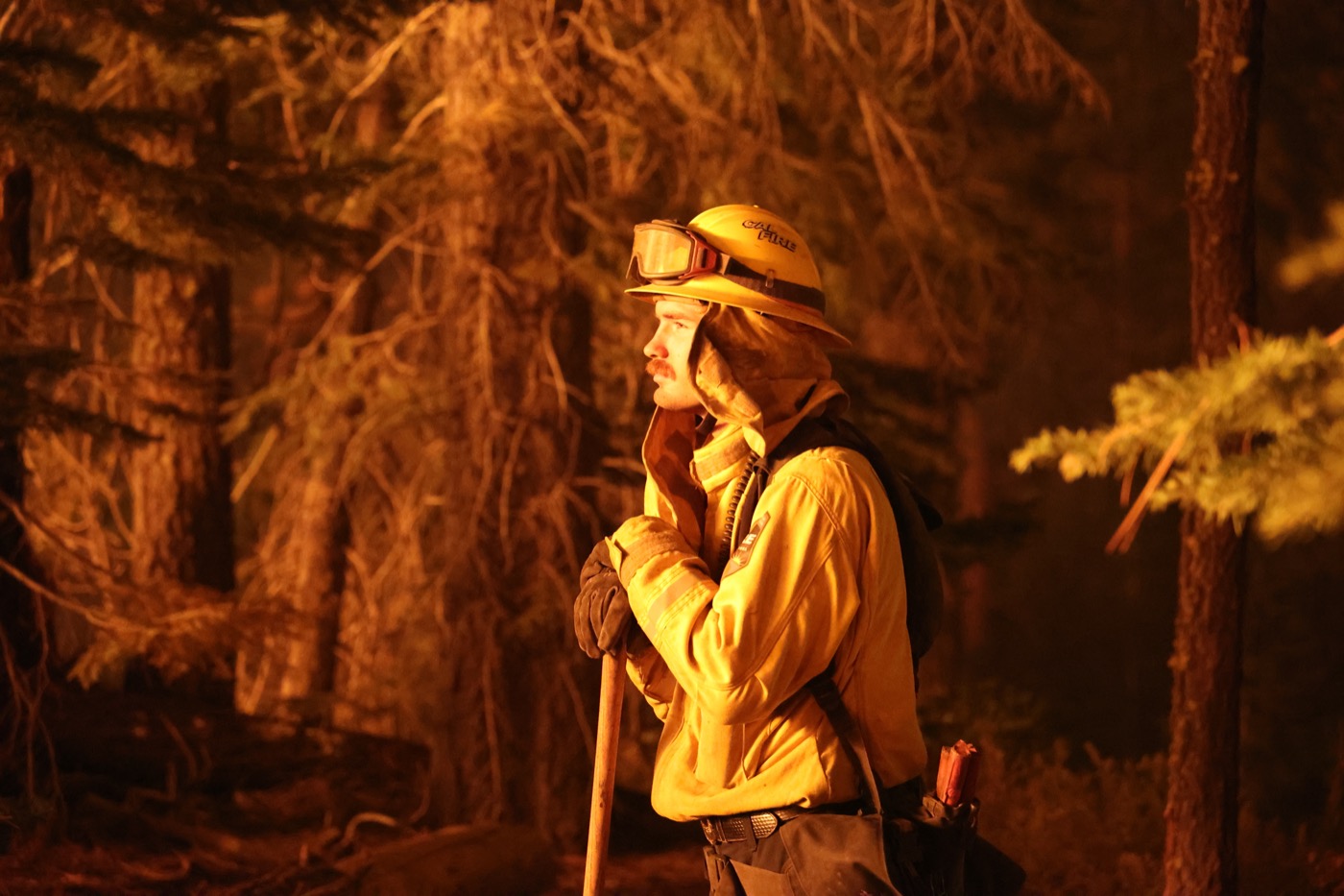 a side profile view of a white man with a moustache wearing a firefighter outfit with a helmet with goggles on it, leaning on a pole. its night and he's being illuminated by the orange glow of the blaze