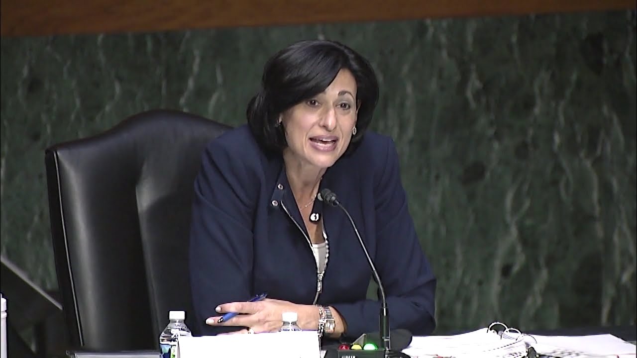 a woman in a suit speaking into a microphone surrounded by binders and water bottles