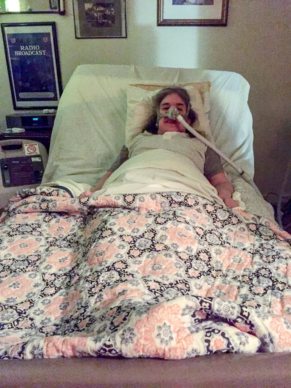 Ingrid is laying in bed with a mask and breathing tube attached to her face. She is looking directly at the camera. There is a pink floral bedspread on the bed and framed posters on the wall behind her.