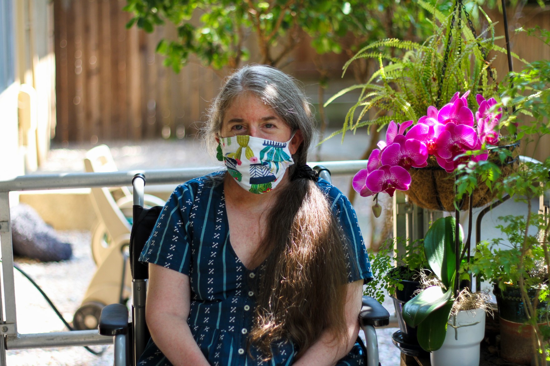 Ingrid, a white woman, is sitting in a chair on her front porch next to a bright magenta orchid. She is in her mid 50's and has gray and brown hair. She's wearing a white face mask with a plant pattern on it, and a patterned blue dress.