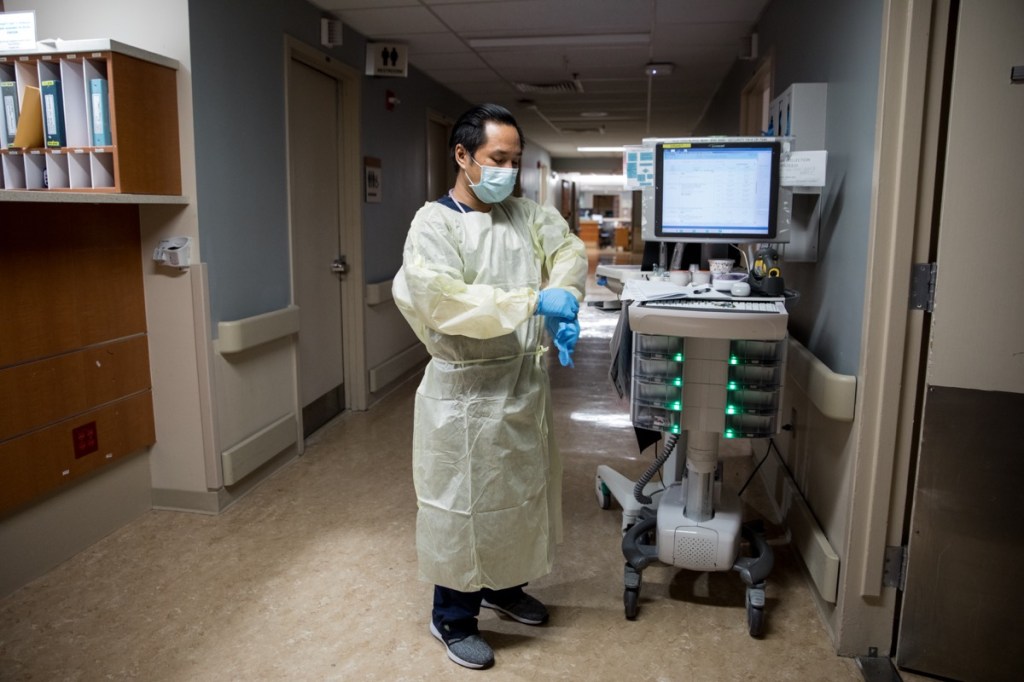 a filipino man in a nursing gown, face mask, and gloves, stands next to a monitor in a hospital hallway