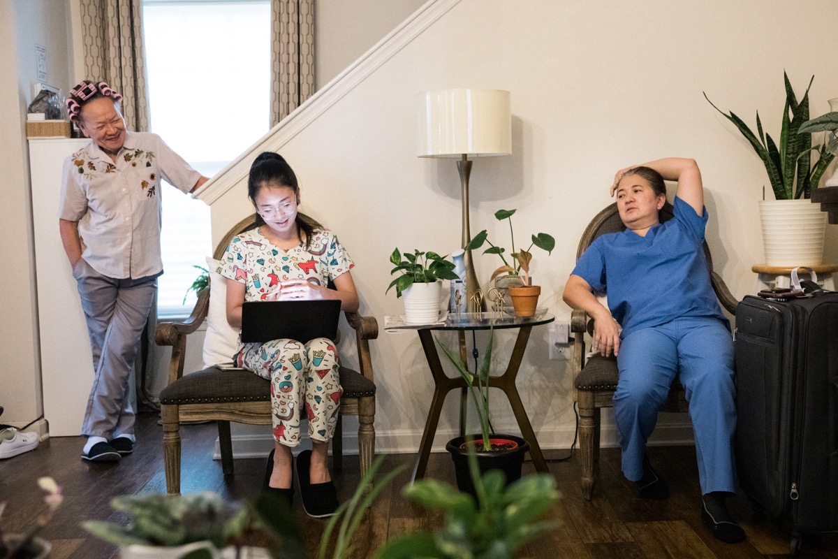 in a room next to a staircase, a filipino woman in a blue nursing uniform sits in a chair with her arm thrown over her head with her daughter also sitting in an arm chair and using a laptop. on the far left standing near the stairwell is an older lady, the daughter's grandmother, with hair curlers smiling as she looks over to her granddaughter