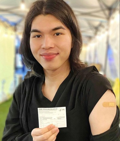 A Filipino nurse in plain clothes smiles at the camera while showing his COVID vaccine card