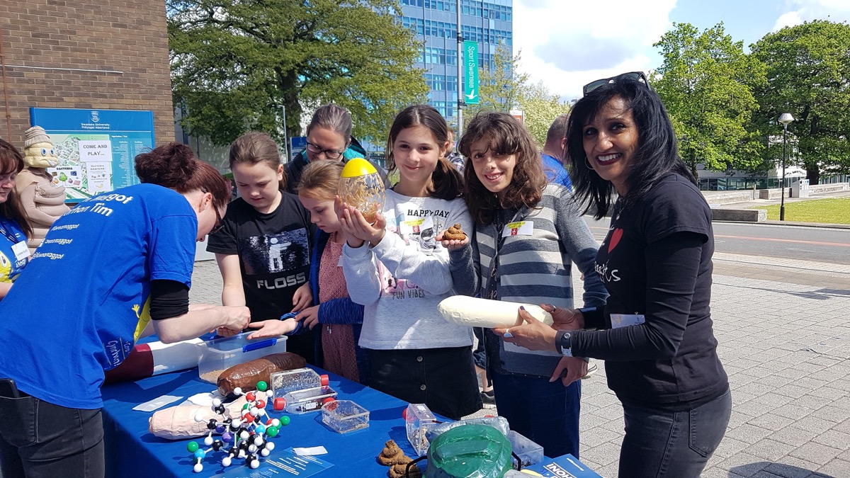 people surrounding a table booth outside. the table is filled with models of insects and maggots. one of the young children holds a large sealed ball of live maggots