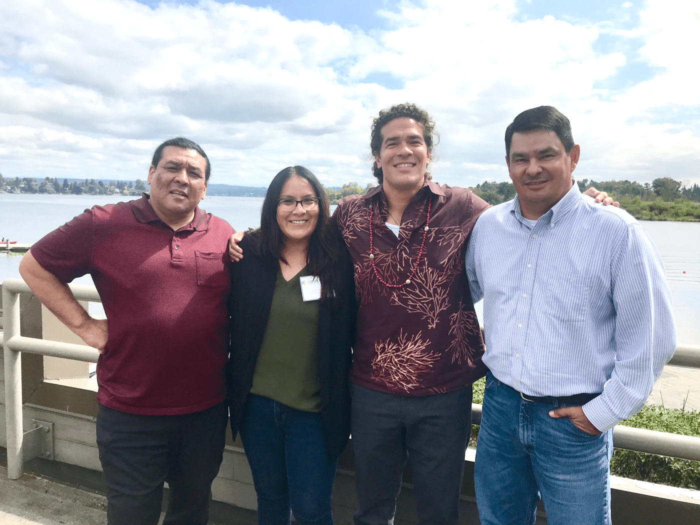 four native americans, three men, one woman, stand next to each other smiling at the camera, with a lake and lush landscape behind them