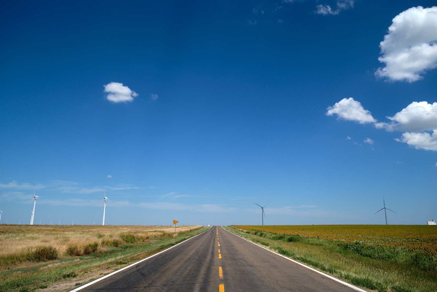 a wide landscape shot take from the middle of a road dividing a large field on a nearly cloudless day. four wind turbines, two on each side of the road, are off in the distance