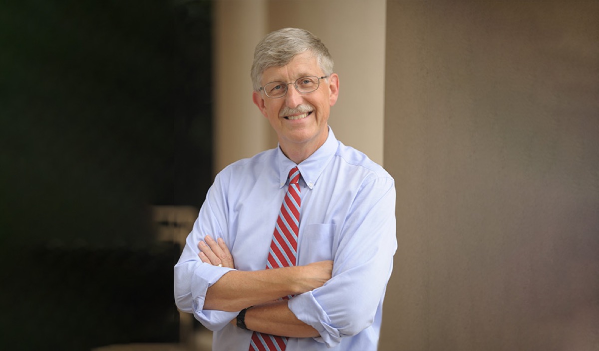 an older white man wearing a button down shirt and striped tie crossing his arms and smiling at the camera