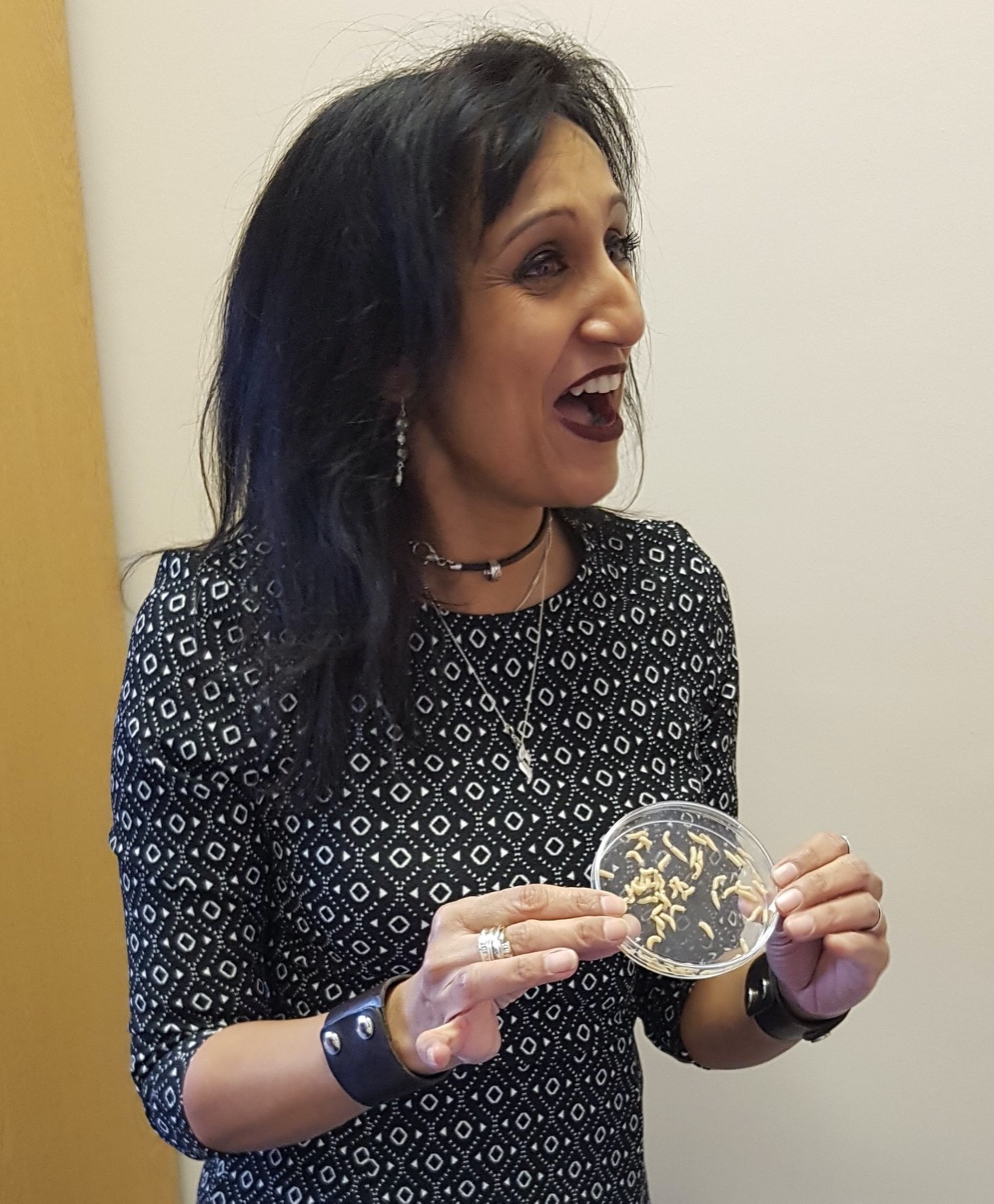 a woman smiling and speaking enthusiastically while holding a petri dish of maggots