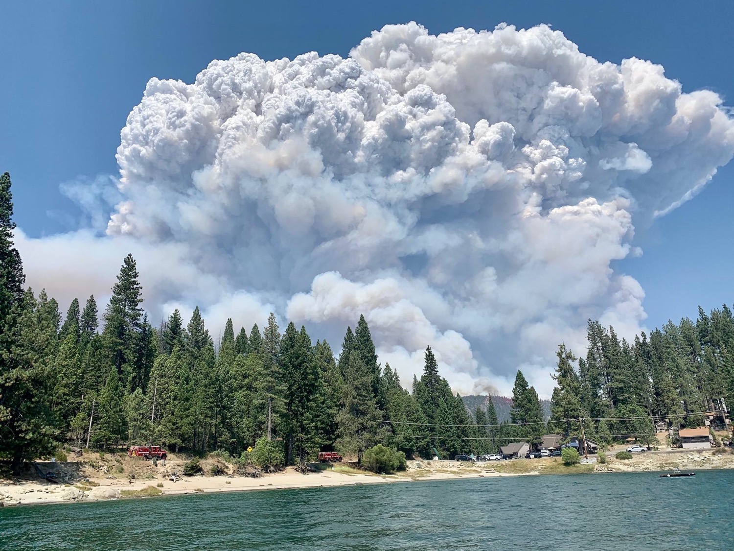 a landscape shot of wilderness with a lake and tall fir trees surround it, and a gigantic plume of smoke obscuring most of the blue sky