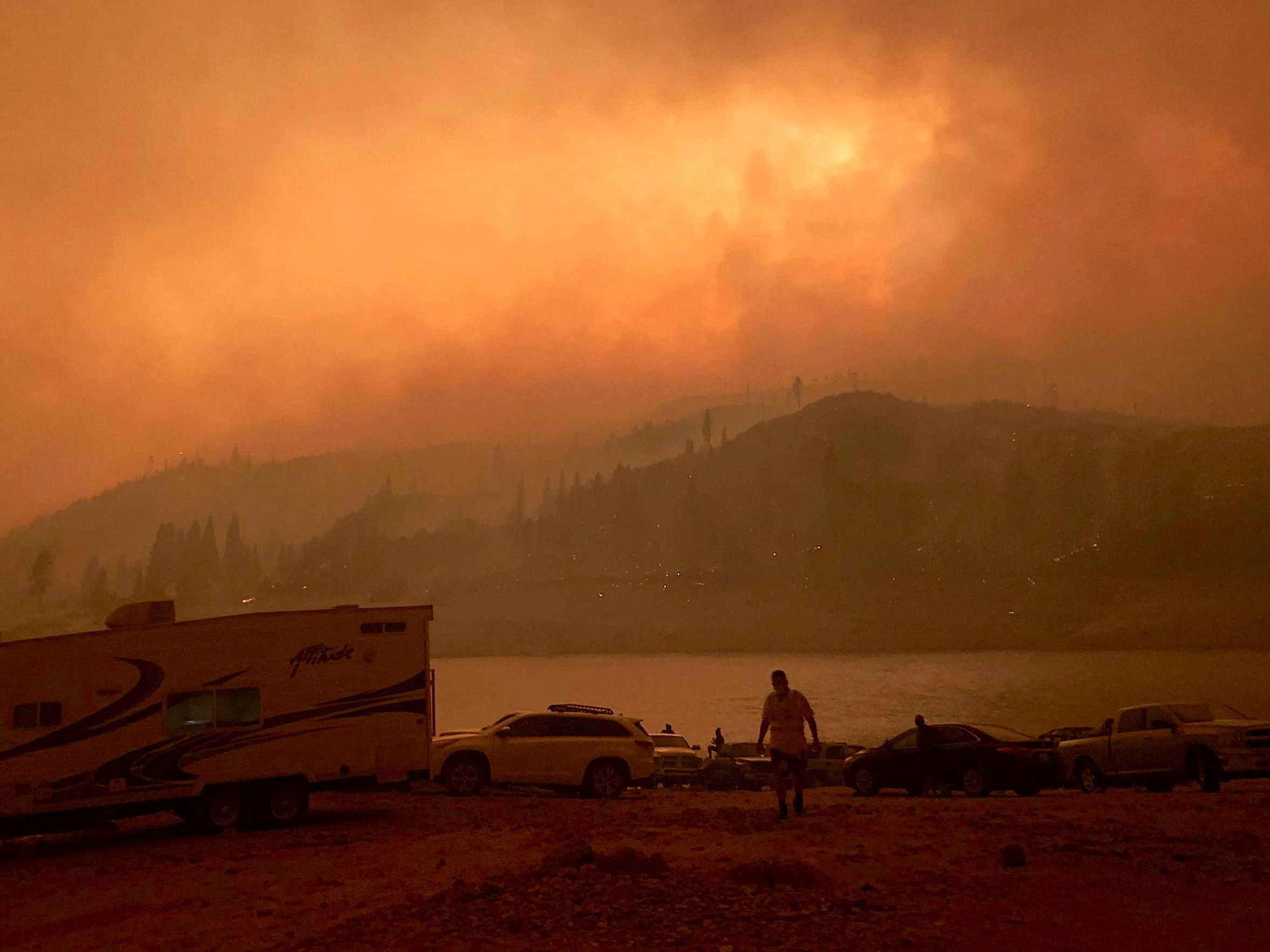 an eery landscape shot of a lake and nearby mountains except the sky is blotted around with thick orange smoke clouds