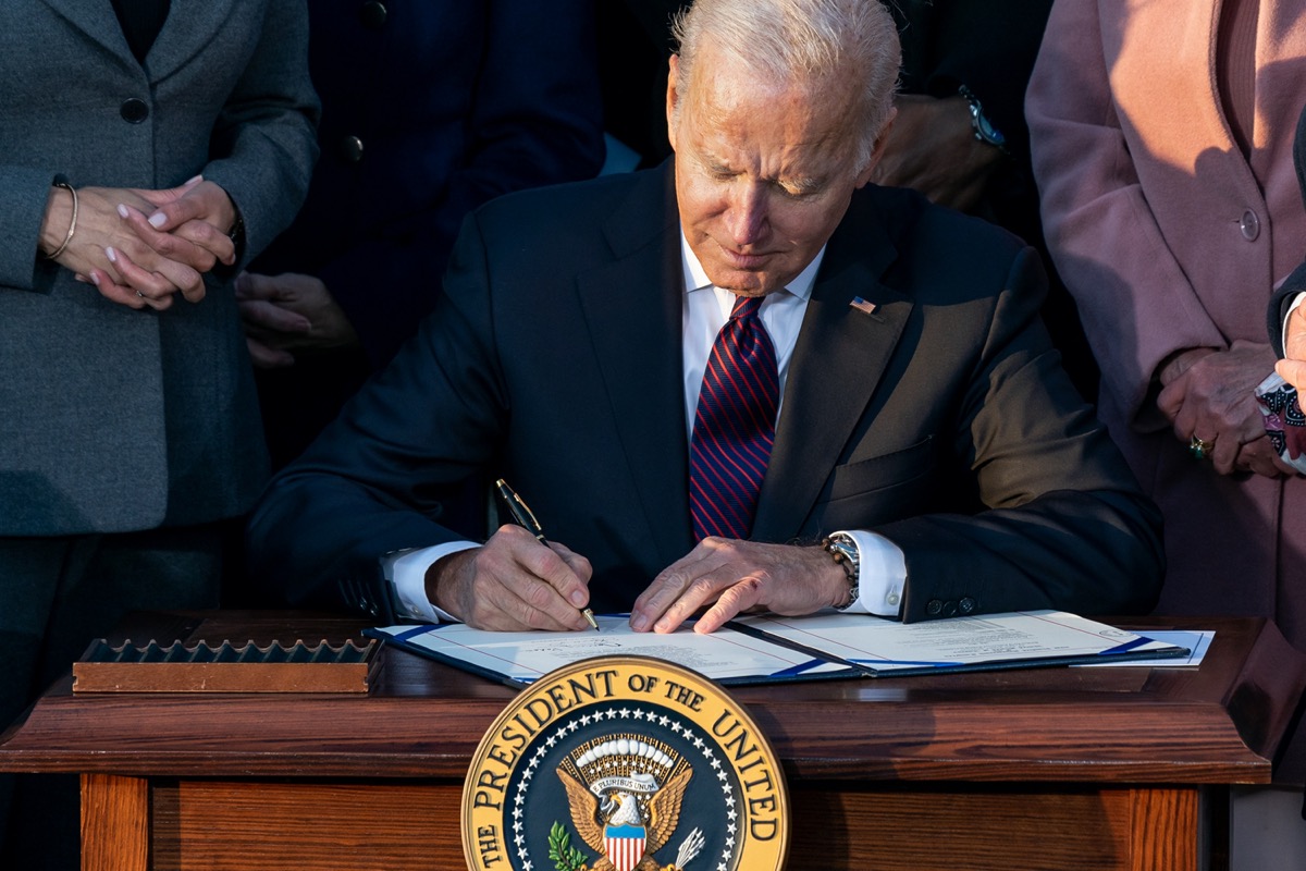a white man with gray hair in suit sits at a desk and signs a piece of paper. in front of the desk is a circular emblem that reads "the president of the united states" and an eagle on it