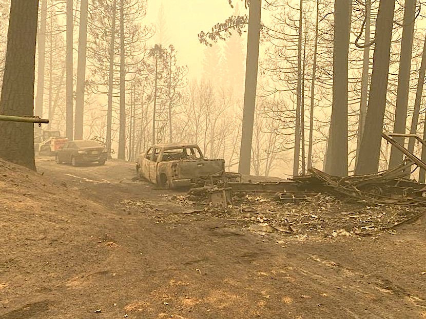 a desolate aftermath of the fire in the forest, featuring hazy grey/orange smoke, no visible vegetation, and the burned husk of a pickup truck