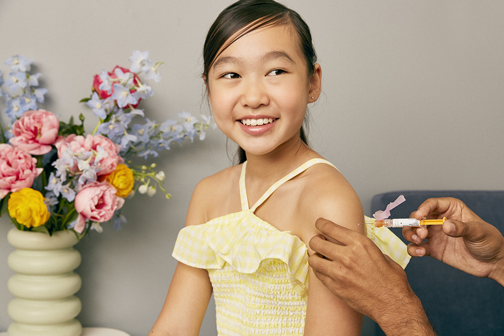 a young asian girl receiving a vaccine shot in her left arm. she's smiling.