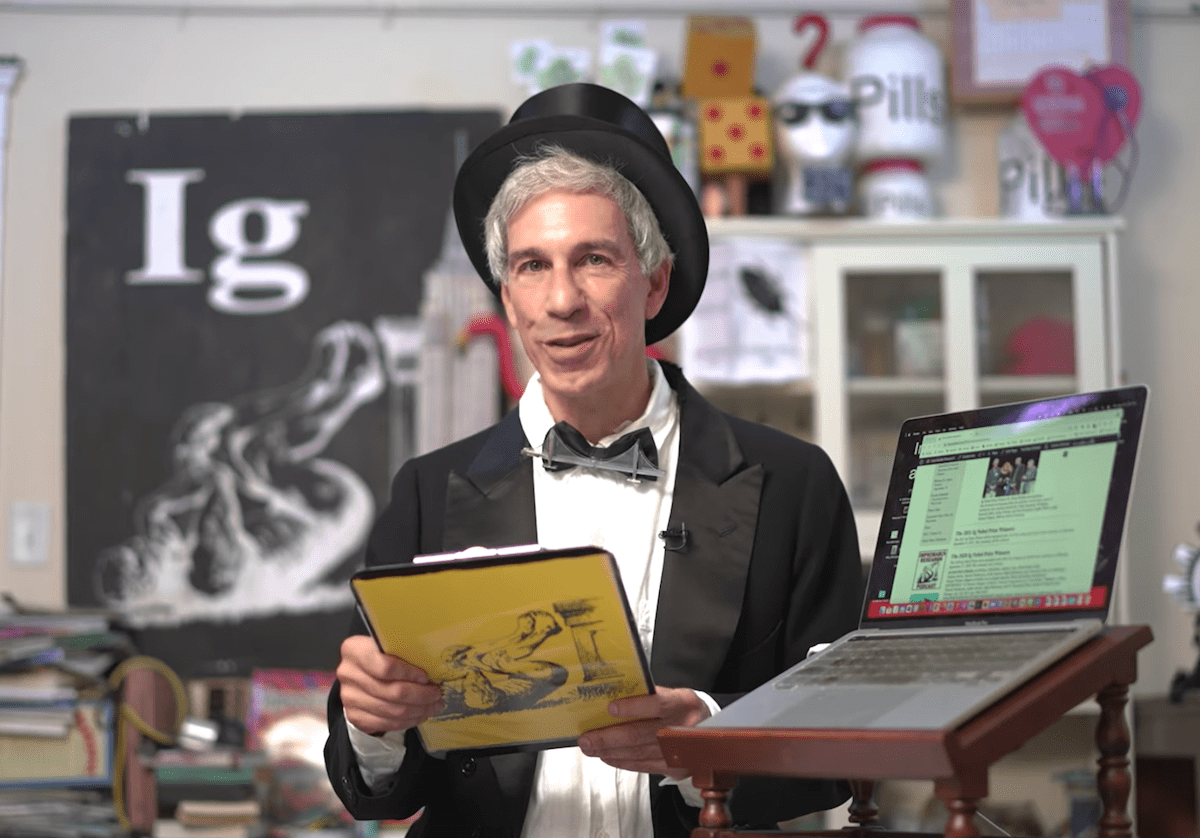 a man with a top hat and a bow tie speaking. there is a laptop next to him and he holds a clipboard. behind him is a black banner that reads ig nobel