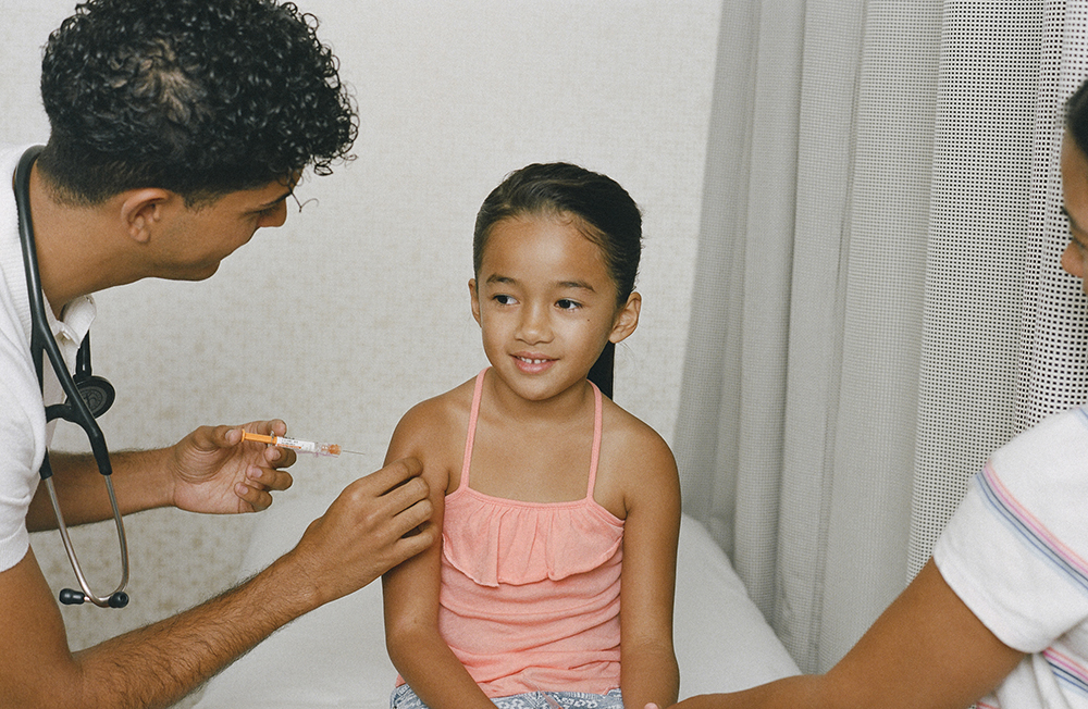 a young girl getting a vaccine shot in her right arm administered by a nurse, with a parent holding her left hand out of frame to her left