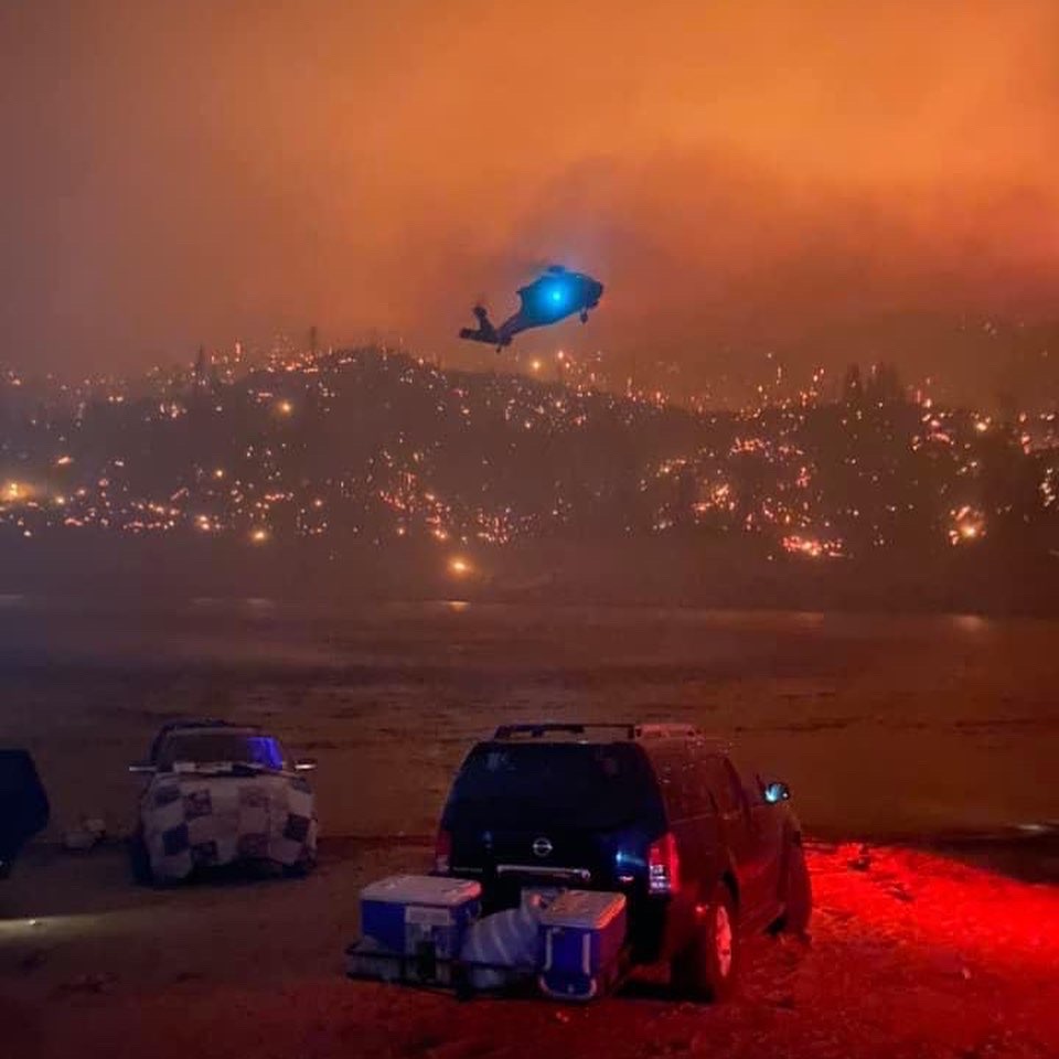 a helicopter with a bright blue searchlight approaches to land at the edge of a lake. its night, but the sky is tinged red from fire and smoke.
