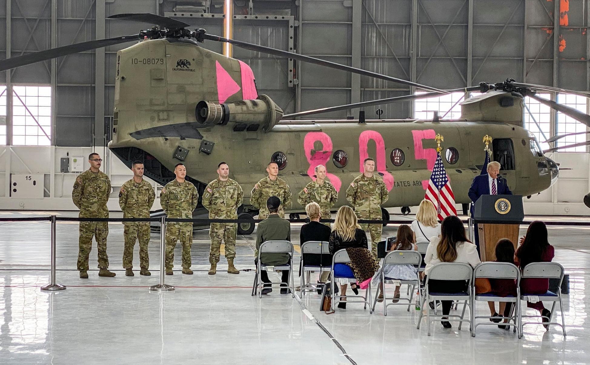 Inside a large hanger, a small crowd looks on as seven members of the california army national guard in fatigues with a large banana helicopter in the background as then president trump stands at a presidential podium giving a speech