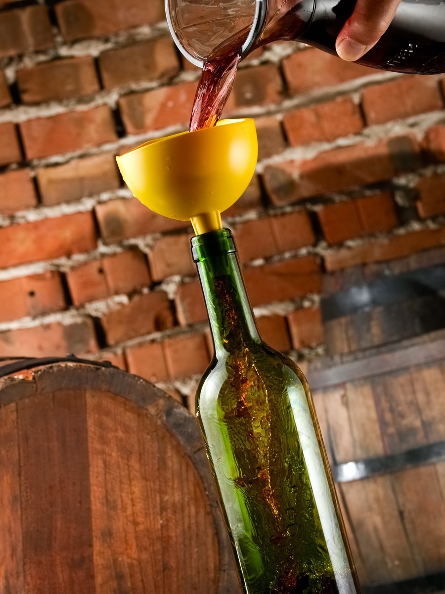 Pouring red wine in a bottle through a yellow funnel, against a background of bricks