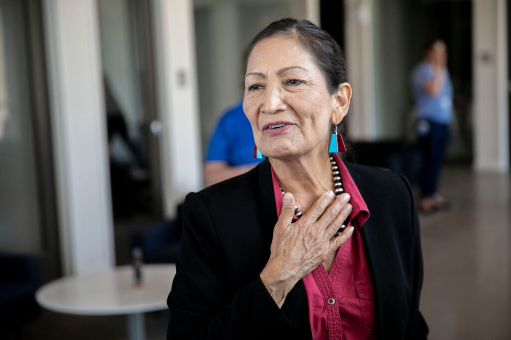 an older indigenous woman with large blue earings smiles with her right hand over the base of her neck, smiling, in a conference room