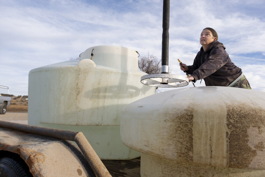 a white woman positions a hose to be lowered into a large white tank, with another large white tank next to it.