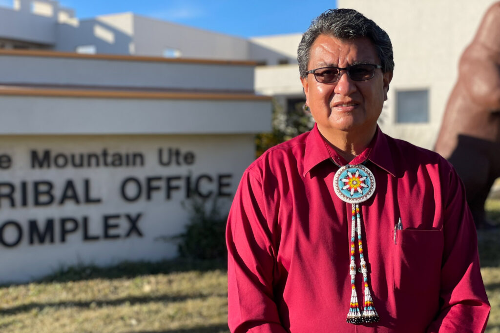 an older indigenous man wearing a red button down shirt and traditional bolo tie and sunglasses stands outside of a building with a sign that reads "tribal office complex"
