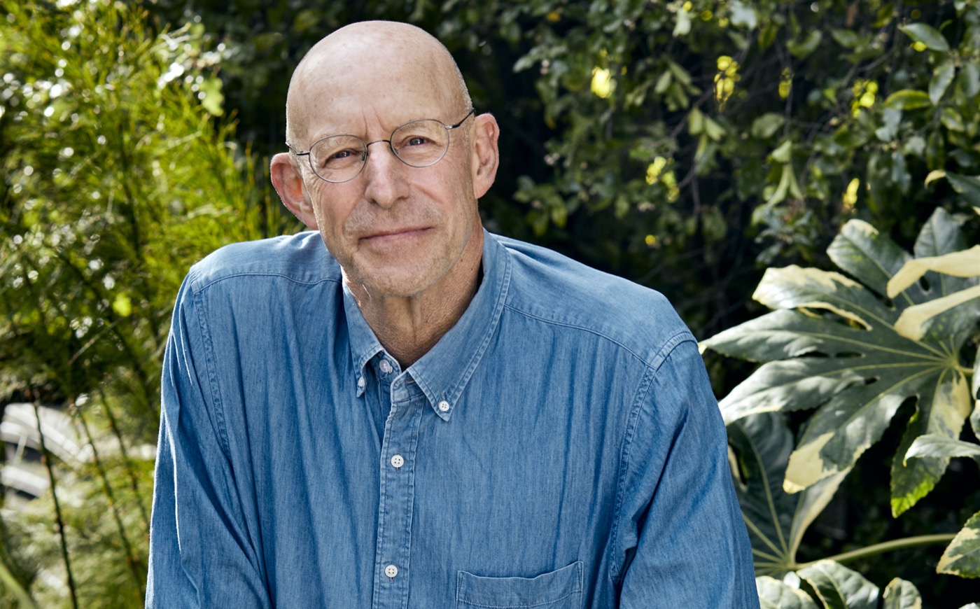 an older white man in a blue button down shirt poses in front of a plant filled background