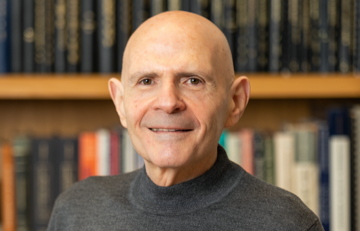 a bald older white man with a bookshelf behind him smiles at the camera