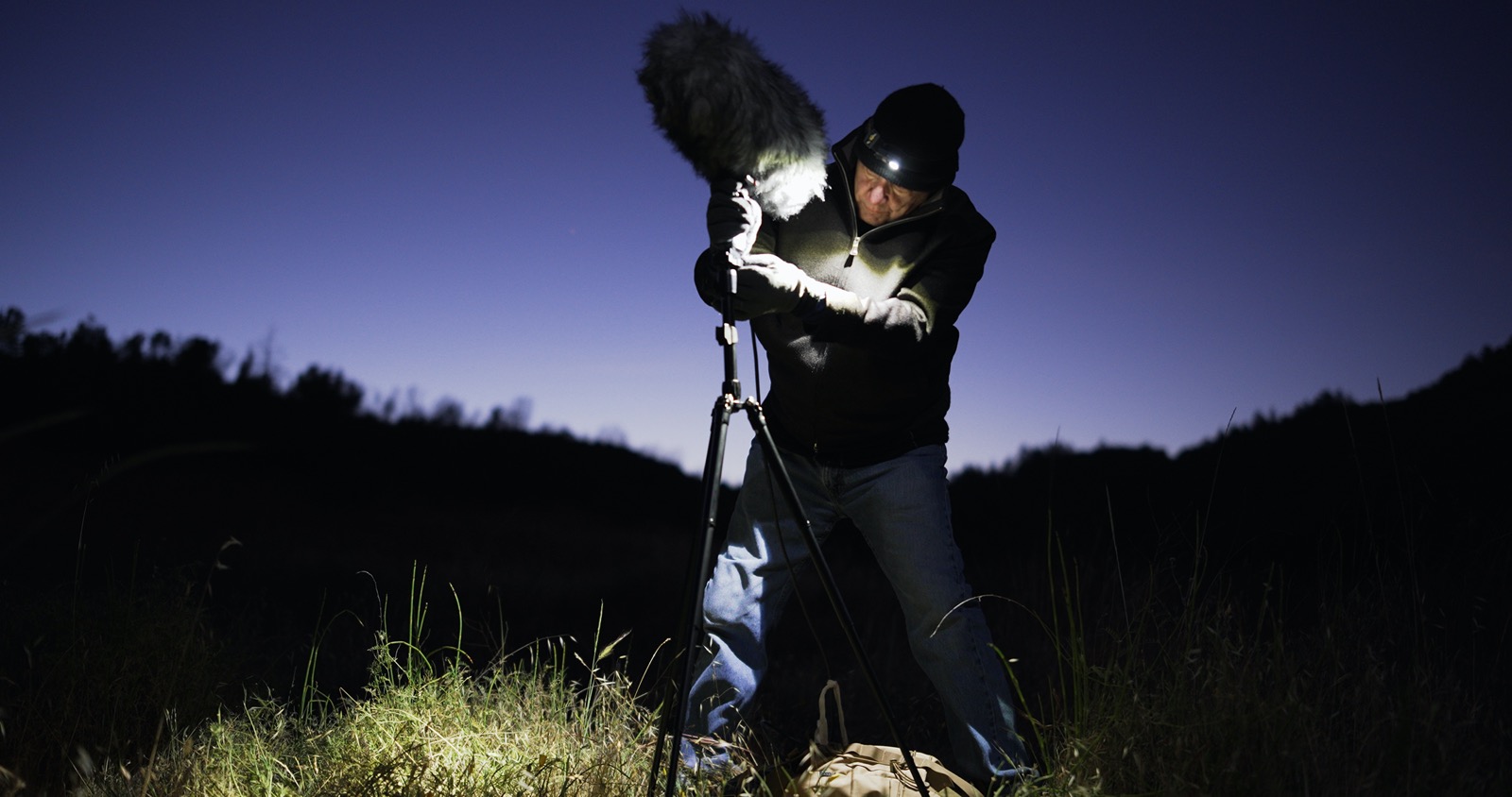 an older white man in cold weather gear wearing a headlamp, sets up a large boom microphone in a field. the sun rises behind him