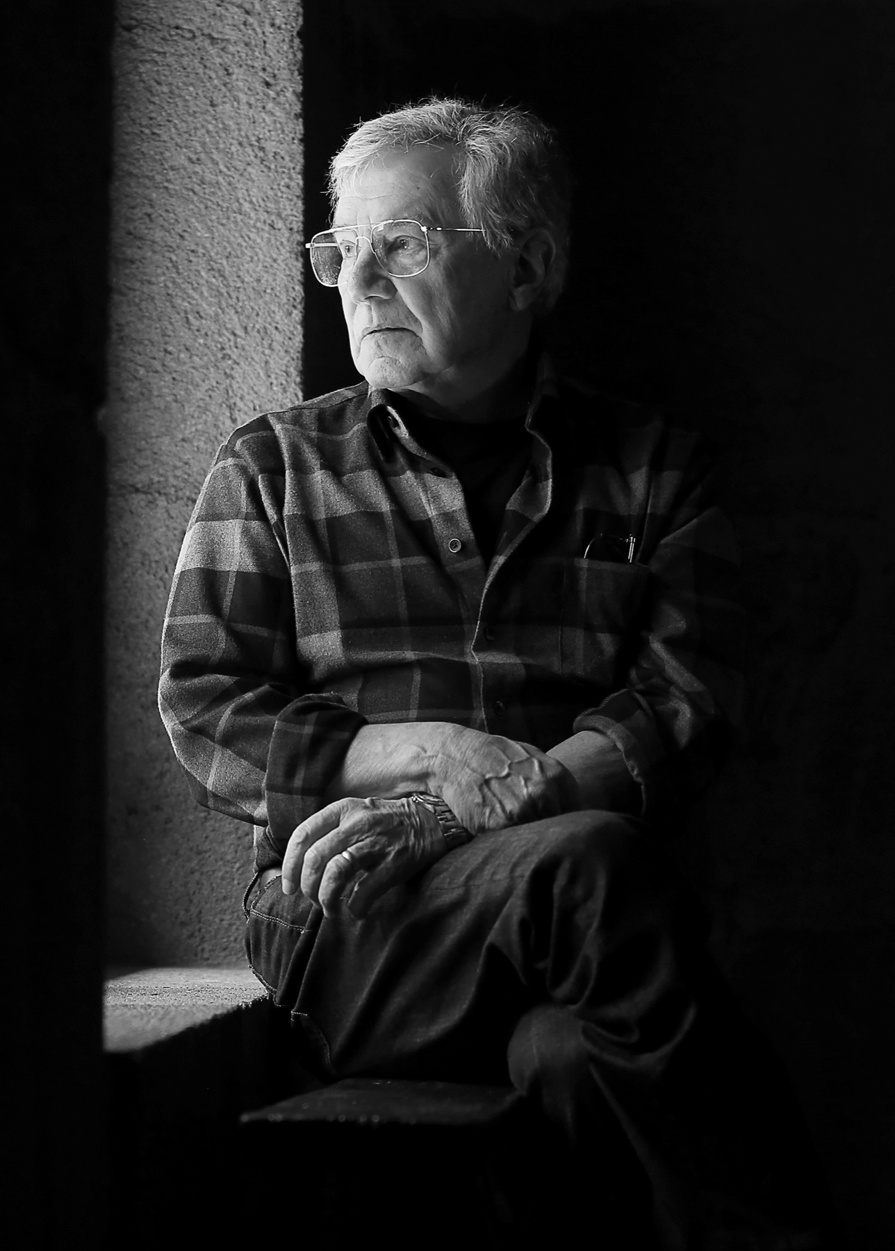 a portrait black and white photo of an older white man looks out from a dark room out to a brighter window