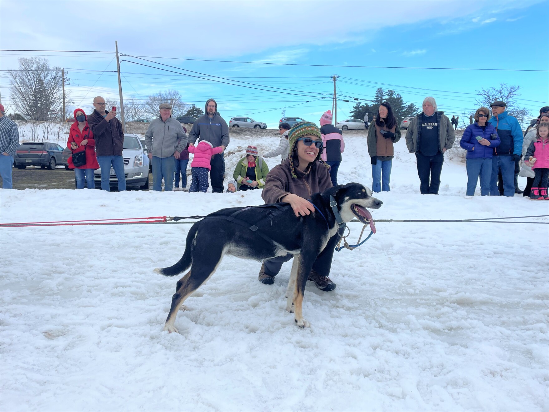 a white woman wearing a beanie and sunglasses and a long sleeve shirt pets a panting dog in front of her. a small crowd stands in the snow behind them