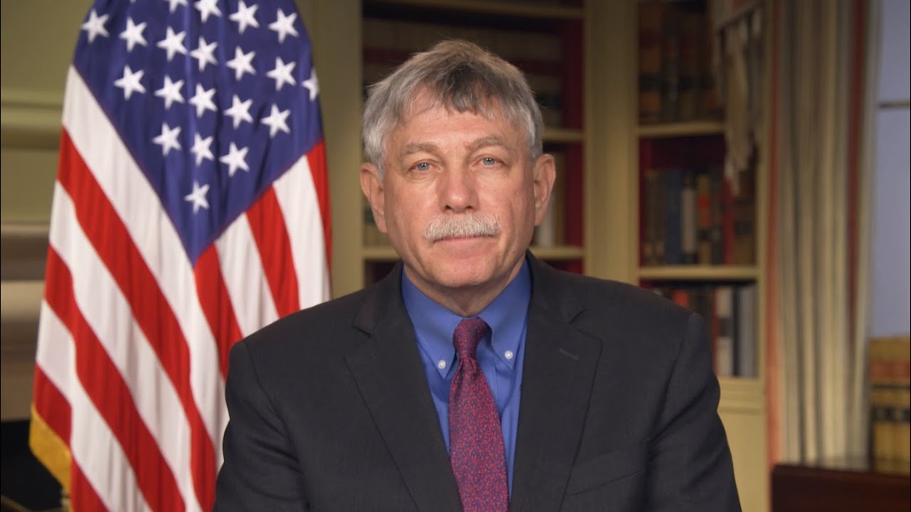 an older white man staring at the camera, with an office and an american flag behind him