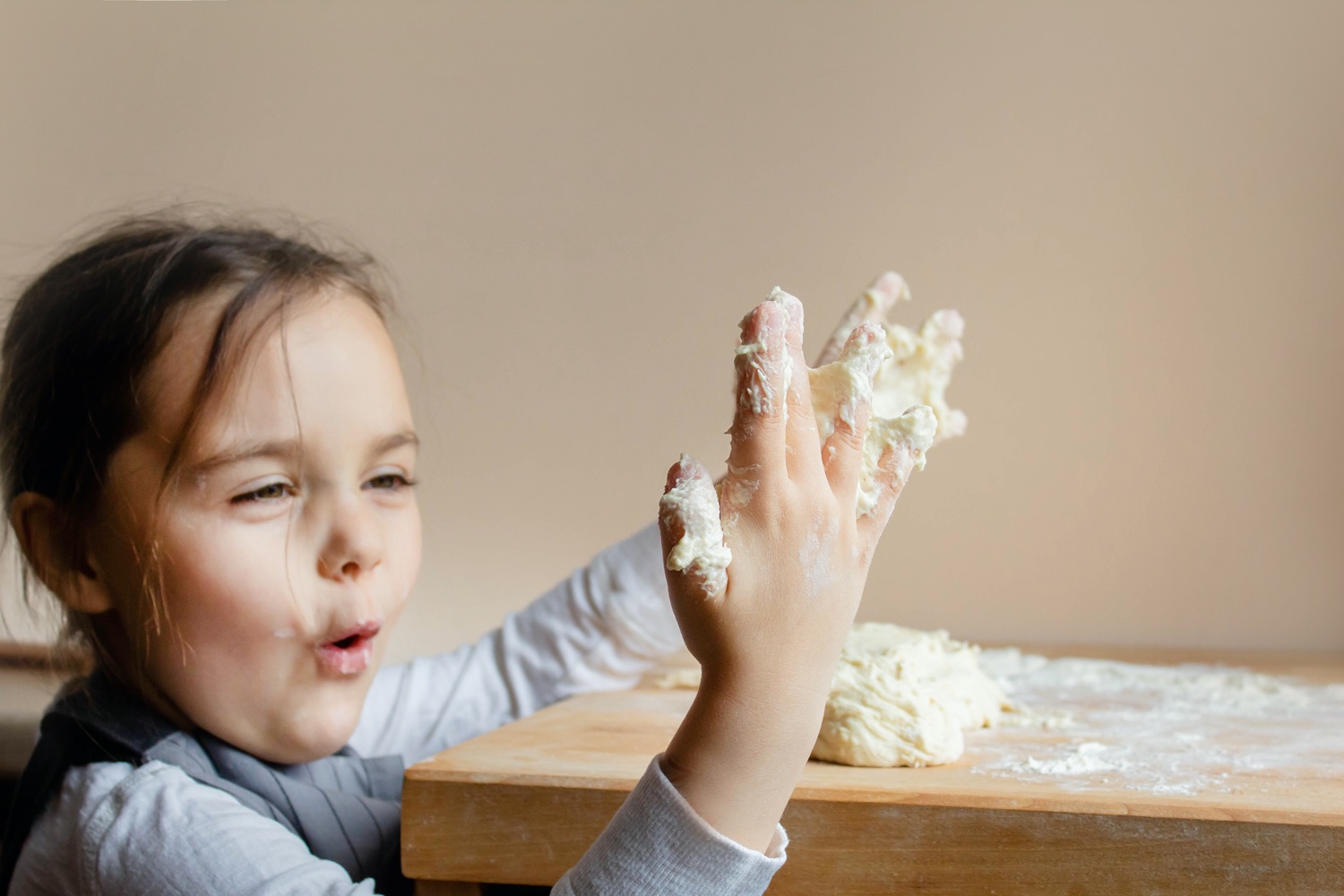 A smiling white little girl dressed as a cook stands at the table and plays with the sticky dough on her fingers in surprise.