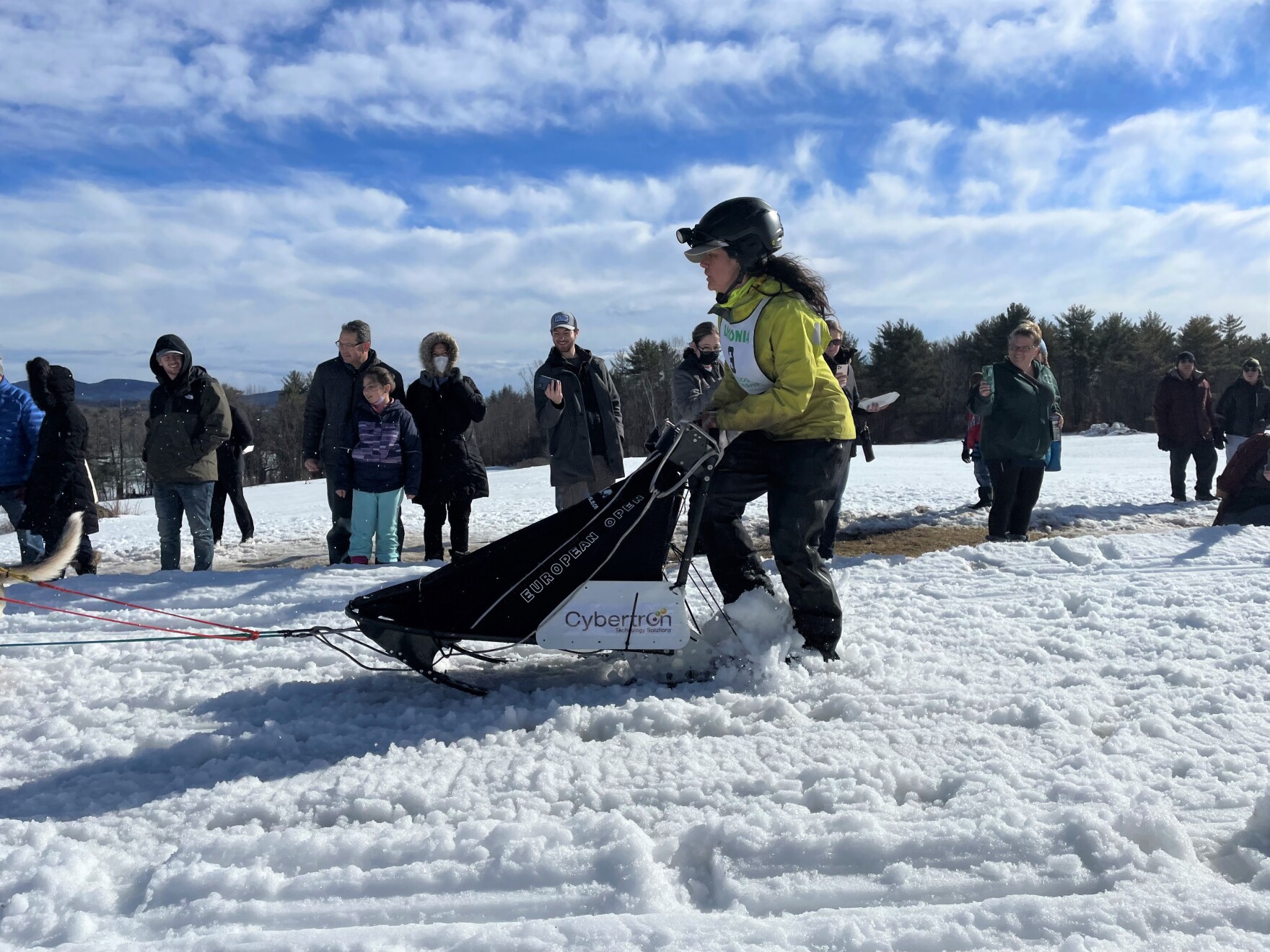 a white woman in snow fear and a helmet stands on a sled in the snow, with a small crowd behind her